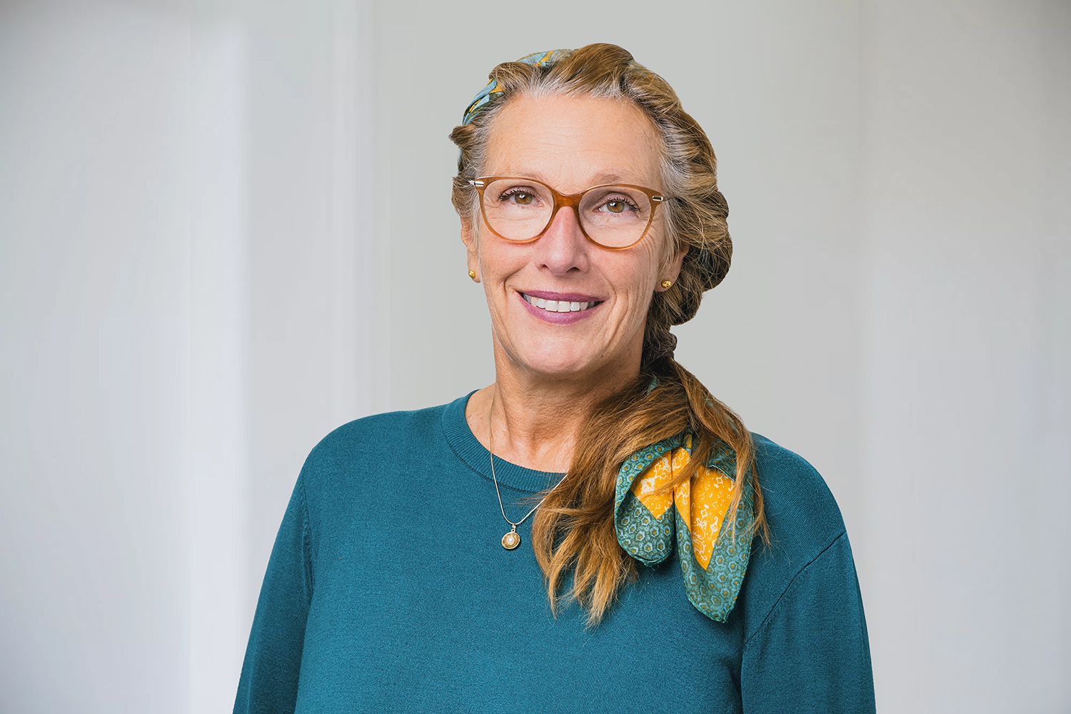 bag-relex_7749 2 Komprimiert eine Frau langen, zusammengebundenen Haaren steht vor einem weißen Hintergrund und lächelt in die Kamera. Sie trägt ein blaues Oberteil und eine Brille.