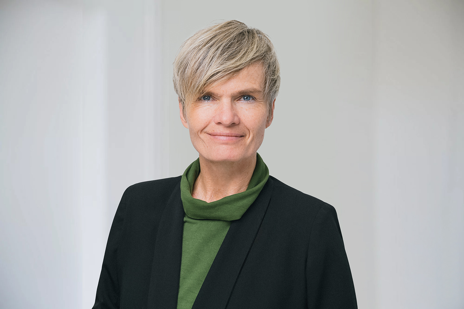 bag-relex_7799 2 Komprimiert eine Frau mit kurzen Haaren steht vor einem weißen Hintergrund und lächelt in die Kamera. Sie trägt einen schwarzen Blazer und ein dunkelgrünes Oberteil darunter.