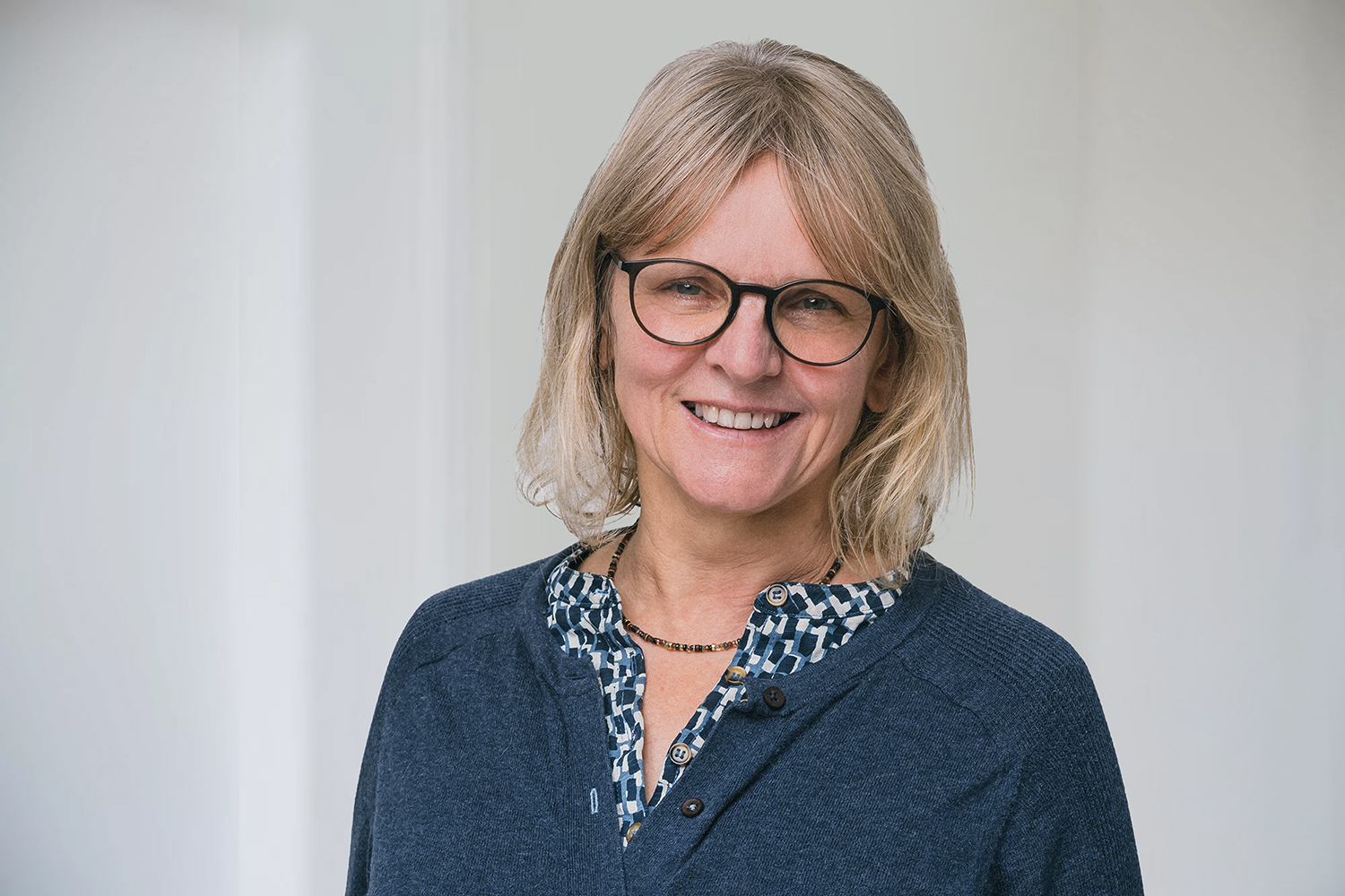 bag-relex_7847 2 Komprimiert eine Frau mit mittellangen Haaren steht vor einem weißen Hintergrund und lächelt in die Kamera. Sie trägt eine Brille und einen dunkelblauen Cadigan mit einer gemusterten Bluse darunter.