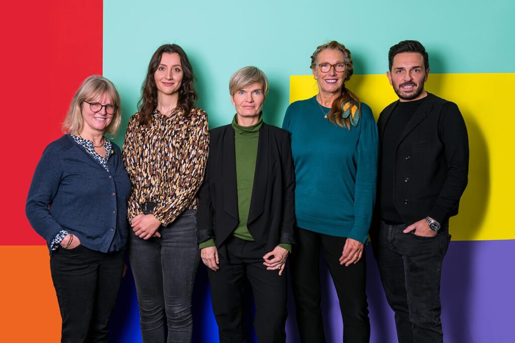 Fünf Menschen stehen vor einem bunten Hintergrund und blicken in die Kamera - Foto des neu gewählten Vorstands der BAG RelEx