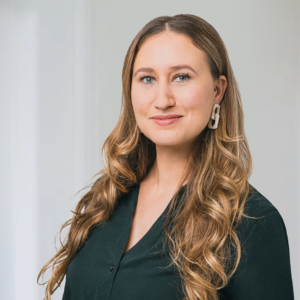 Eine Frau mit langen Haaren steht vor einem weißen Hintergrund und lächelt in die Kamera. Sie trägt ein schwarzes Oberteil.