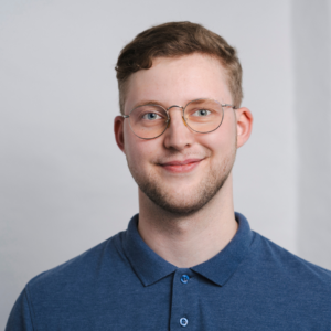 Dehne_BAG RelEx Ein Mann mit kurzen Haaren und Brille steht vor einem weißen Hintergrund und blickt lächelnd in die Kamera. Er trägt ein blaues Oberteil.